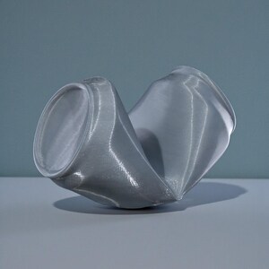 May include: A crushed, gray aluminum can, resting on a gray surface against a blue-gray backdrop. The can is severely dented, with its cylindrical shape distorted and flattened. The top and bottom rims are still visible.