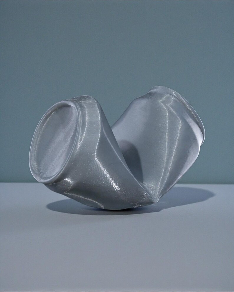 May include: A crushed, gray aluminum can, resting on a gray surface against a blue-gray backdrop. The can is severely dented, with its cylindrical shape distorted and flattened. The top and bottom rims are still visible.
