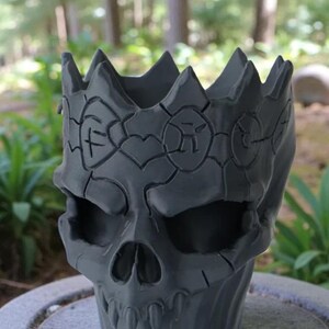May include: A gray skull-shaped planter with a crown design. The planter has a detailed, textured surface and is suitable for small plants or decorative items. The skull features eye sockets, a nose cavity, and teeth.