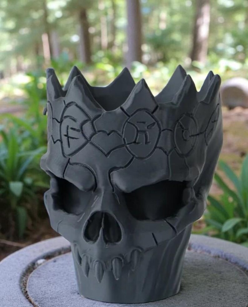 May include: A gray skull-shaped planter with a crown design. The planter has a detailed, textured surface and is suitable for small plants or decorative items. The skull features eye sockets, a nose cavity, and teeth.