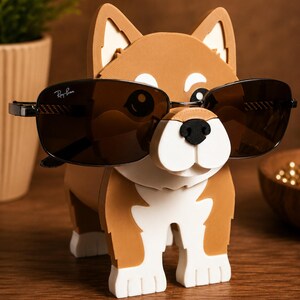 May include: A brown and white dog-shaped eyeglass holder with black sunglasses. The holder has a cartoonish design with a white snout and paws. The sunglasses are black with the Ray-Ban logo.