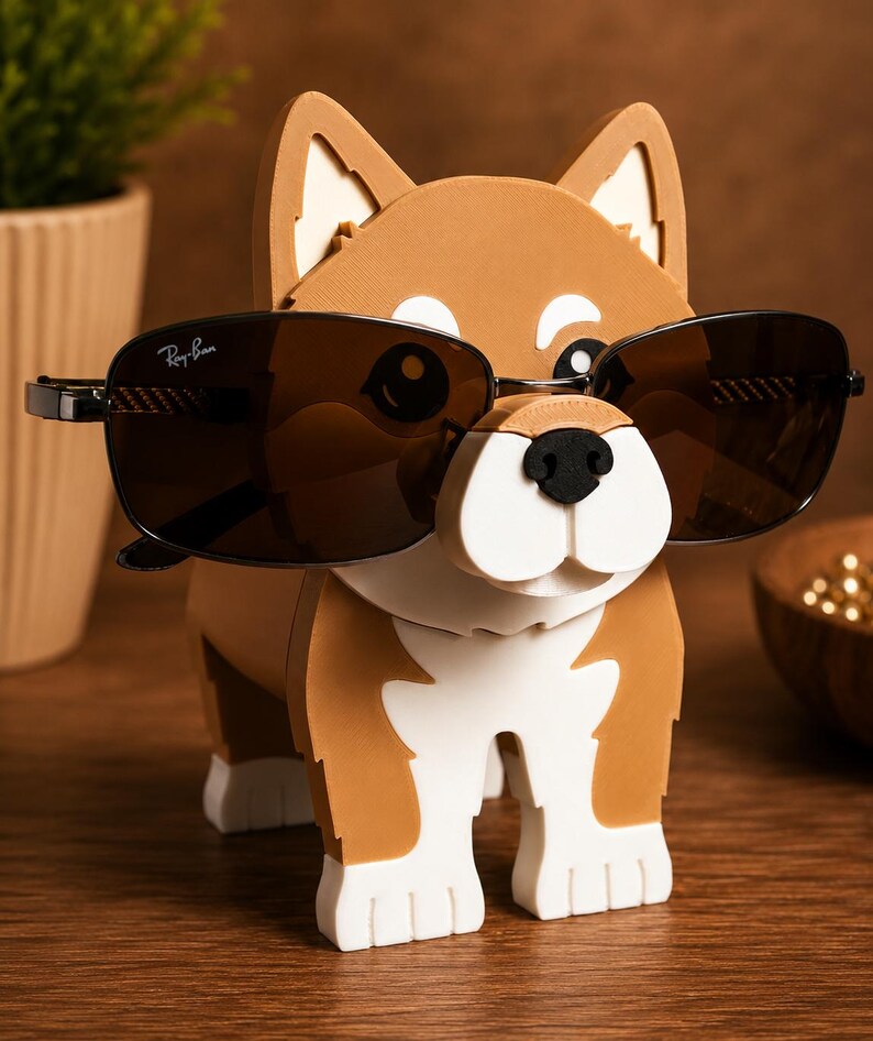 May include: A brown and white dog-shaped eyeglass holder with black sunglasses. The holder has a cartoonish design with a white snout and paws. The sunglasses are black with the Ray-Ban logo.