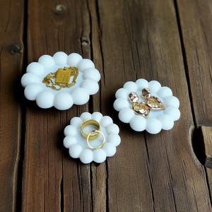 May include: Three white, flower-shaped jewelry dishes on a wooden surface. Each dish holds different gold jewelry: a necklace, earrings, and rings. The dishes have a textured, bubble-like design.