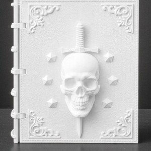 Peut inclure: Un livre blanc textur&eacute; avec un design de cr&acirc;ne et d'&eacute;p&eacute;e. Le cr&acirc;ne est centr&eacute;, avec une &eacute;p&eacute;e qui le transperce. Des accents d&eacute;coratifs d'angle et des formes g&eacute;om&eacute;triques entourent le cr&acirc;ne. Le livre a une tranche avec des charni&egrave;res.