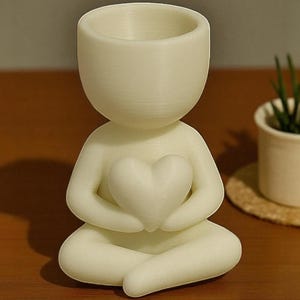 May include: A cream-colored, seated figure holding a heart, designed as a small planter. The figure has a large head with a hollowed-out top for planting. The figure is in a meditative pose, with legs crossed and arms holding the heart.
