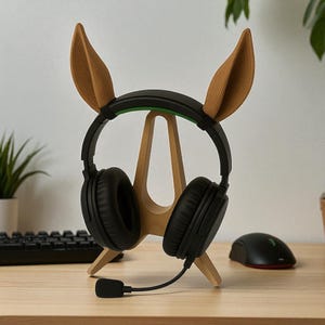 Puede incluir: Un soporte de auriculares de madera con orejas de conejo marrones que sostiene unos auriculares negros con micr&oacute;fono. El soporte est&aacute; sobre un escritorio de madera, con un teclado y un rat&oacute;n en el fondo.