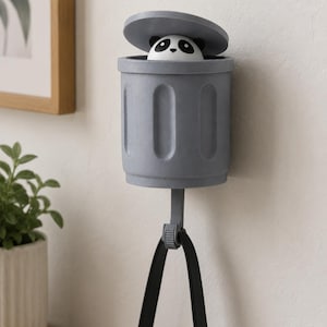 May include: A gray wall-mounted hook shaped like a trash can, with a panda bear peeking out from inside. The hook has a black strap hanging from it, and is mounted on a white wall.