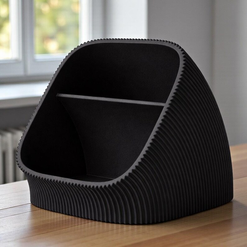 May include: A black desk tidy with a distinctive, curved form. The organiser has a ribbed texture on the outside and a horizontal divider inside, creating two compartments. The overall shape resembles a stylized arch.