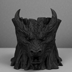May include: A black, stylized planter shaped like a mythical beast's head. The planter features a detailed, textured surface with horns, a snout, and an open mouth. The design resembles a carved tree trunk.