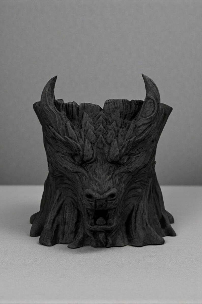 May include: A black, stylized planter shaped like a mythical beast's head. The planter features a detailed, textured surface with horns, a snout, and an open mouth. The design resembles a carved tree trunk.