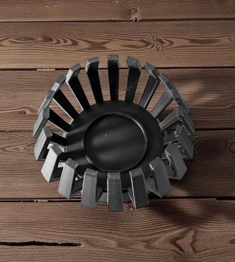 Puede incluir: Un cuenco decorativo de metal negro con base circular y soportes rectangulares verticales. El cuenco se encuentra sobre un fondo de madera, creando un contraste de texturas y tonos. El dise&ntilde;o es moderno y geom&eacute;trico.