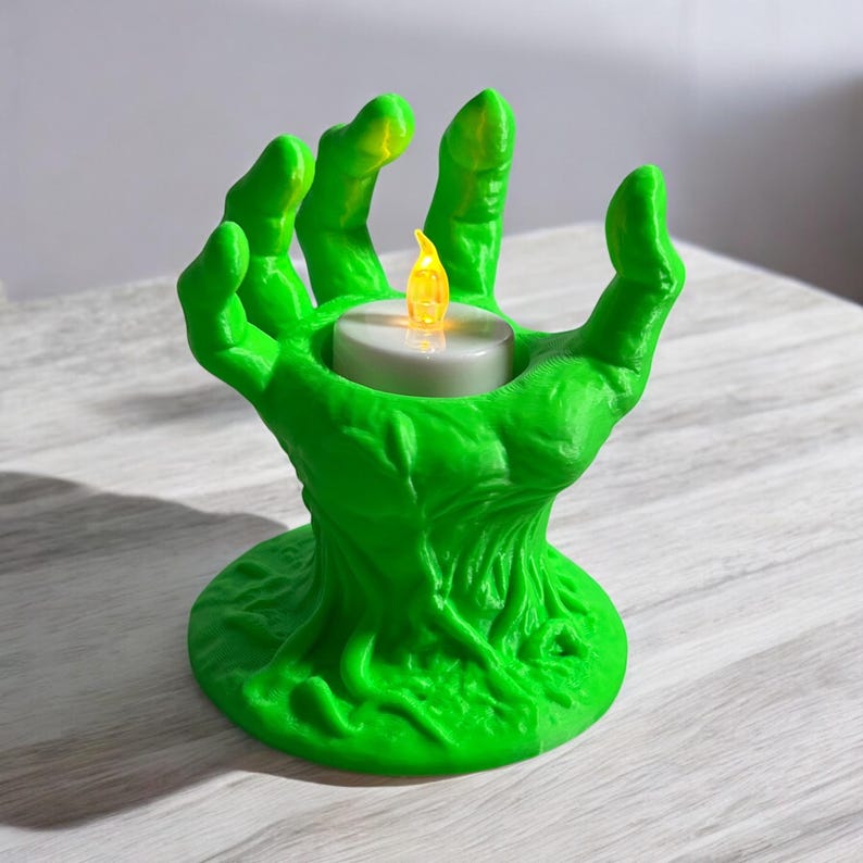 May include: A green, 3D-printed candle holder shaped like a hand. The hand is holding a small, white candle with a flickering yellow flame. The candle holder has a textured surface and is sitting on a light-colored surface.