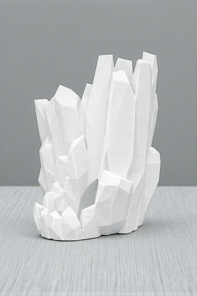 May include: A white, geometric crystal sculpture with a cluster of sharp, angular forms. The sculpture has a textured surface and is set against a neutral gray background. The design resembles a geode or a cluster of crystals.