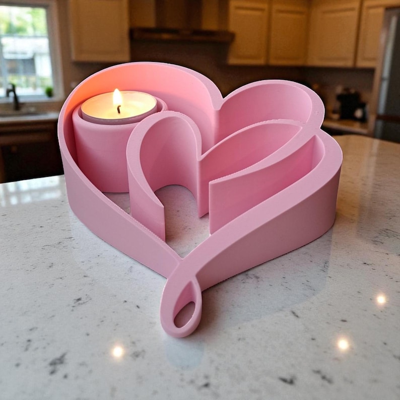 May include: A pink heart-shaped candle holder with a lit candle. The holder has a unique design with curved lines and a loop at the bottom. The candle holder is made of a solid material and sits on a countertop.