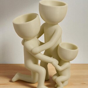 Op de afbeelding: Een cr&egrave;mekleurig familiebeeldje dat een knielende figuur, een volwassen figuur en een kinderfiguur uitbeeldt. De figuren zijn verbonden, waarbij de volwassen figuren het kind omarmen. Het beeldje is gemaakt van een glad materiaal en staat op een houten oppervlak.