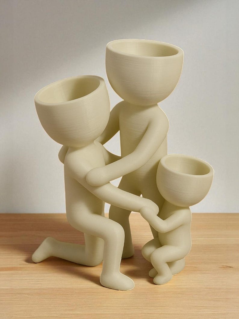 Op de afbeelding: Een cr&egrave;mekleurig familiebeeldje dat een knielende figuur, een volwassen figuur en een kinderfiguur uitbeeldt. De figuren zijn verbonden, waarbij de volwassen figuren het kind omarmen. Het beeldje is gemaakt van een glad materiaal en staat op een houten oppervlak.