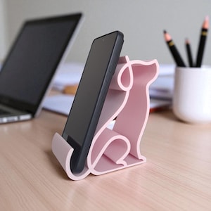 May include: A pink dog-shaped phone stand holding a black smartphone. The stand is made of a solid material and has a minimalist design. The phone is angled slightly, and the stand is on a wooden desk. A laptop and pencils are in the background.