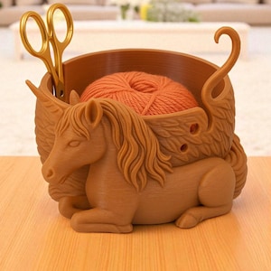 Pode incluir: Uma tigela de fio castanha, impressa em 3D, em forma de unicórnio reclinado. A tigela contém uma bola de fio laranja e tesouras douradas. O design apresenta uma crina detalhada, asas e uma alça curva. Ideal para tricotar e fazer crochê.