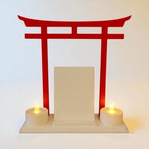May include: A red Torii gate-shaped display with a white base, a central rectangular panel, and two battery-operated candle lights. The gate is a traditional Japanese architectural structure.