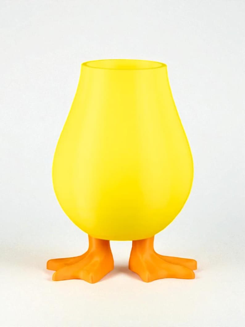 Puede incluir: Un jarr&oacute;n amarillo caprichoso con forma de pato, con un cuerpo redondeado y una parte superior abierta. El jarr&oacute;n tiene patas de pato naranjas como base, creando un art&iacute;culo decorativo l&uacute;dico y &uacute;nico. El color amarillo brillante es llamativo.