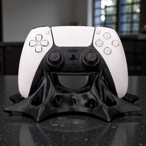 May include: A white and black PlayStation 5 controller resting on a black, stylized stand. The controller has a white top and black bottom with buttons and joysticks. The stand has a unique, angular design.