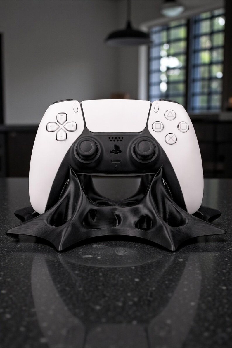 May include: A white and black PlayStation 5 controller resting on a black, stylized stand. The controller has a white top and black bottom with buttons and joysticks. The stand has a unique, angular design.