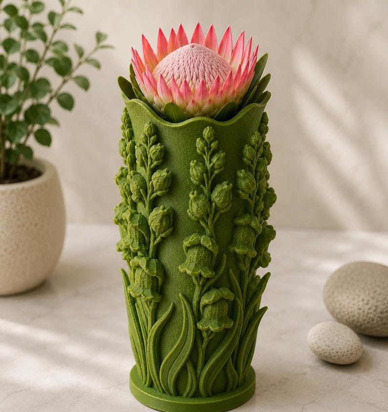 May include: A green vase with a detailed floral design, holding a large pink and red protea flower. The vase has a textured surface with sculpted lily of the valley and leaves. The flower is in full bloom, with a soft pink center and vibrant red-tipped petals.
