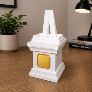May include: A white decorative pedestal with a gold-colored plaque. The pedestal has a stepped design and a central, inverted V-shaped element. The item is displayed on a wooden surface.