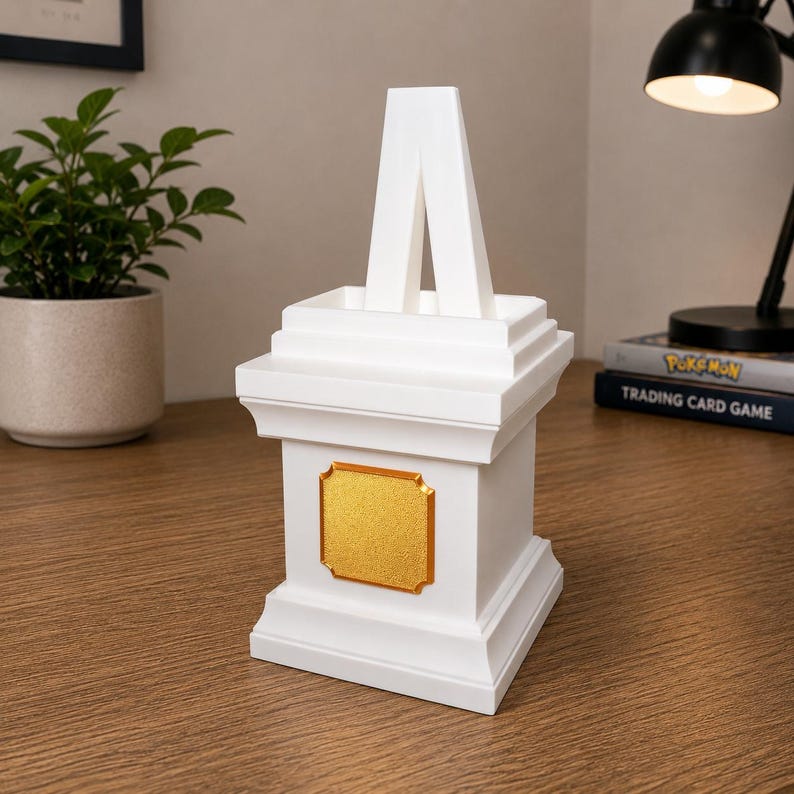 May include: A white decorative pedestal with a gold-colored plaque. The pedestal has a stepped design and a central, inverted V-shaped element. The item is displayed on a wooden surface.