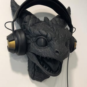 May include: A black dragon head sculpture wearing headphones. The dragon features textured scales, horns, and an open mouth. The headphones are black with gold accents. A black cord hangs down.