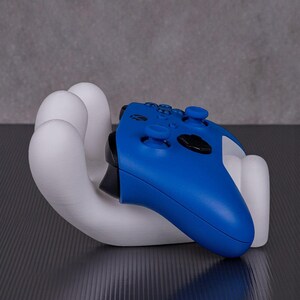 May include: A white, hand-shaped gaming controller holder cradles a blue controller. The controller has black accents and buttons. The holder is on a dark surface with a gray background.