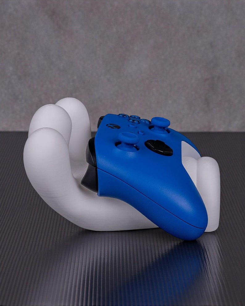 May include: A white, hand-shaped gaming controller holder cradles a blue controller. The controller has black accents and buttons. The holder is on a dark surface with a gray background.