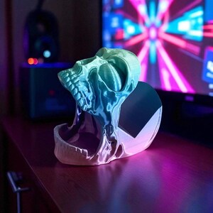 Pu&ograve; includere: Un supporto per telefono a forma di teschio stampato in 3D nei toni del blu e del verde. Il teschio &egrave; dettagliato con denti e orbite visibili e il supporto ha un retro piatto. L'oggetto &egrave; appoggiato su una superficie di legno, con uno schermo luminoso sullo sfondo.