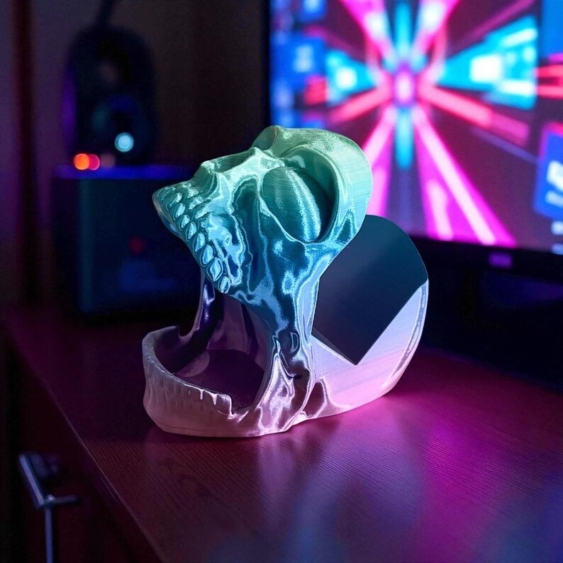 Pu&ograve; includere: Un supporto per telefono a forma di teschio stampato in 3D nei toni del blu e del verde. Il teschio &egrave; dettagliato con denti e orbite visibili e il supporto ha un retro piatto. L'oggetto &egrave; appoggiato su una superficie di legno, con uno schermo luminoso sullo sfondo.