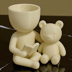 May include: A cream-coloured decorative planter shaped like a person reading a book, with a teddy bear beside them. The planter has a large, open head for planting. The figures are made of a smooth, solid material.