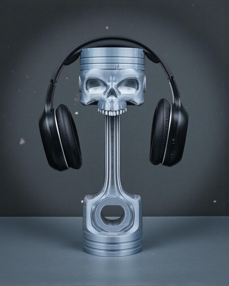 May include: A silver-colored, skull-shaped piston with a set of black headphones resting on top. The piston is a unique decorative item, possibly a headphone stand, with a detailed skull design and a mechanical aesthetic.