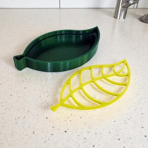 May include: Two leaf-shaped kitchen accessories. One is a solid, dark green dish. The other is a yellow, openwork leaf-shaped item. Both are made of a plastic material and are designed to resemble leaves.