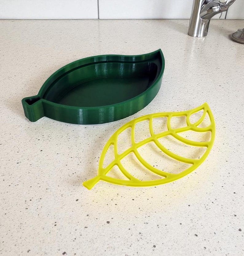 May include: Two leaf-shaped kitchen accessories. One is a solid, dark green dish. The other is a yellow, openwork leaf-shaped item. Both are made of a plastic material and are designed to resemble leaves.