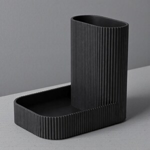 May include: A black, ribbed desk organizer with a tall, oval-shaped pen holder and a rectangular tray. The organizer is made of a matte material and sits on a light gray surface. The design is minimalist and modern.