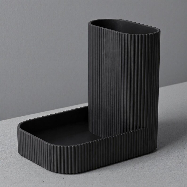 May include: A black, ribbed desk organizer with a tall, oval-shaped pen holder and a rectangular tray. The organizer is made of a matte material and sits on a light gray surface. The design is minimalist and modern.