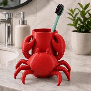 May include: A red crab-shaped toothbrush holder with a light blue toothbrush. The holder has a cylindrical cup with crab claws and a crab body. The holder is on a marble countertop in a bathroom setting.