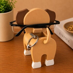 Peut inclure: Un organiseur de bureau fantaisiste en forme de chien, de couleur beige et marron, avec des accents blancs sur les pieds. L'organisateur contient une paire de lunettes &agrave; monture noire et un jeu de cl&eacute;s. Une petite plante en pot et un livre sont en arri&egrave;re-plan.