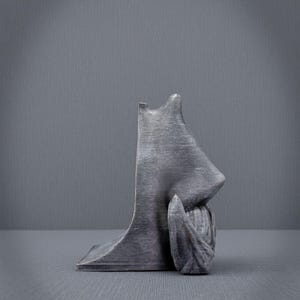 May include: A gray sculpture of a human profile, featuring a nose and a hand resting near the face. The sculpture has a textured surface and is set against a gray backdrop. The artwork is a unique decorative piece.