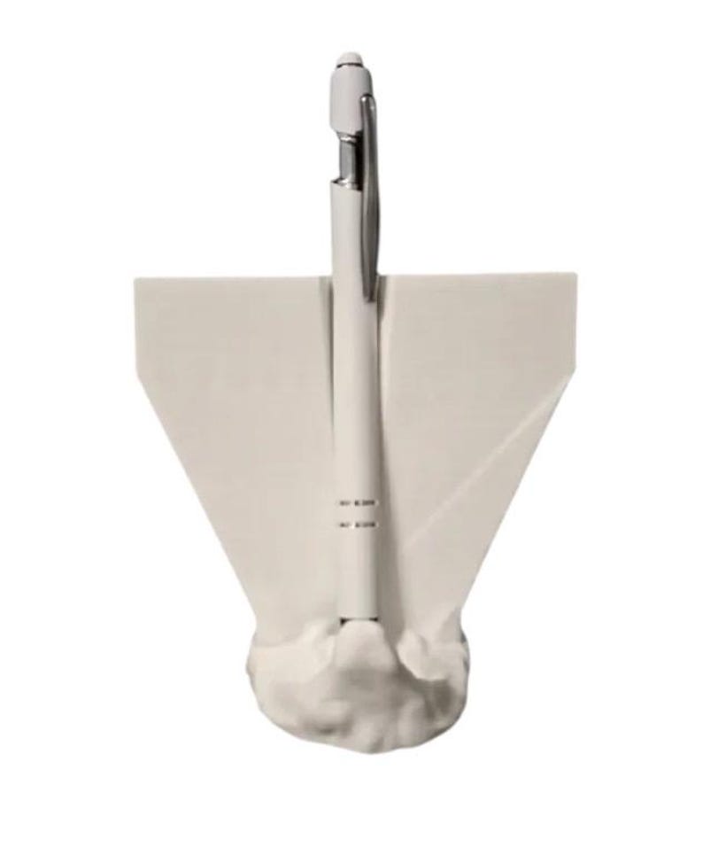 May include: A white pen with a silver clip is displayed in a white pen holder. The pen holder has a unique, abstract design with a wide base and angled sides, creating a modern aesthetic. The pen is centered in the holder.