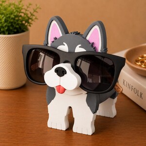 May include: A whimsical dog-shaped eyeglass holder in shades of gray, white, and black, with pink inner ears and a red tongue. The holder is wearing black sunglasses. A small potted plant and a book are in the background.