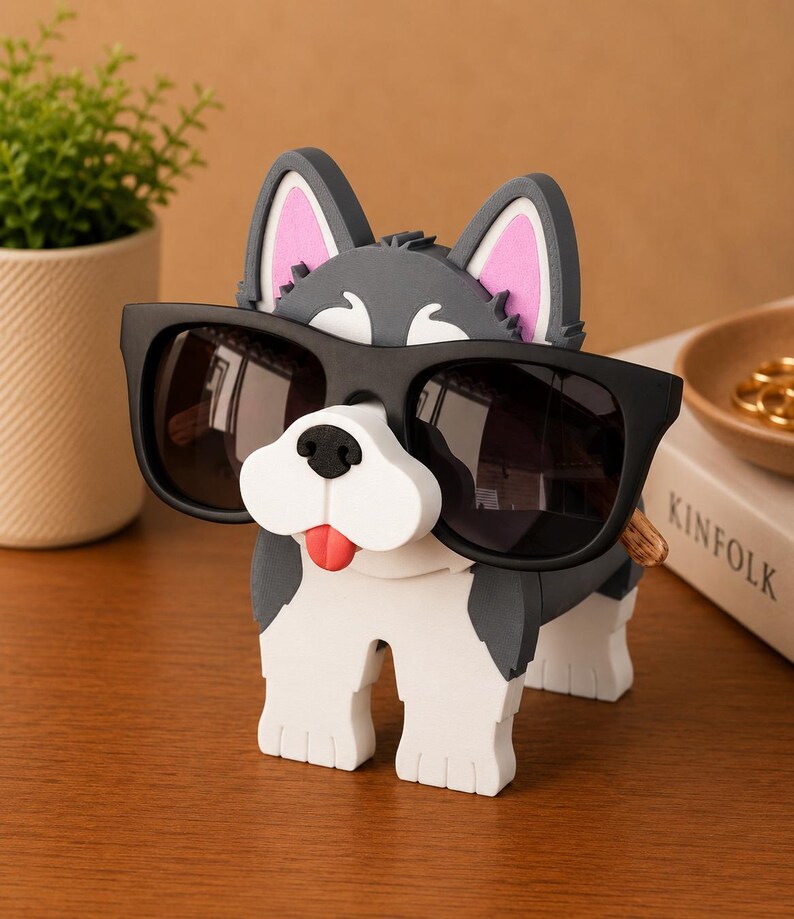 May include: A whimsical dog-shaped eyeglass holder in shades of gray, white, and black, with pink inner ears and a red tongue. The holder is wearing black sunglasses. A small potted plant and a book are in the background.