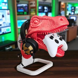May include: A red dinosaur-shaped gaming headset and controller stand. The stand holds a black headset with gold accents and a white controller. The stand is on a white base and sits on a wooden surface.