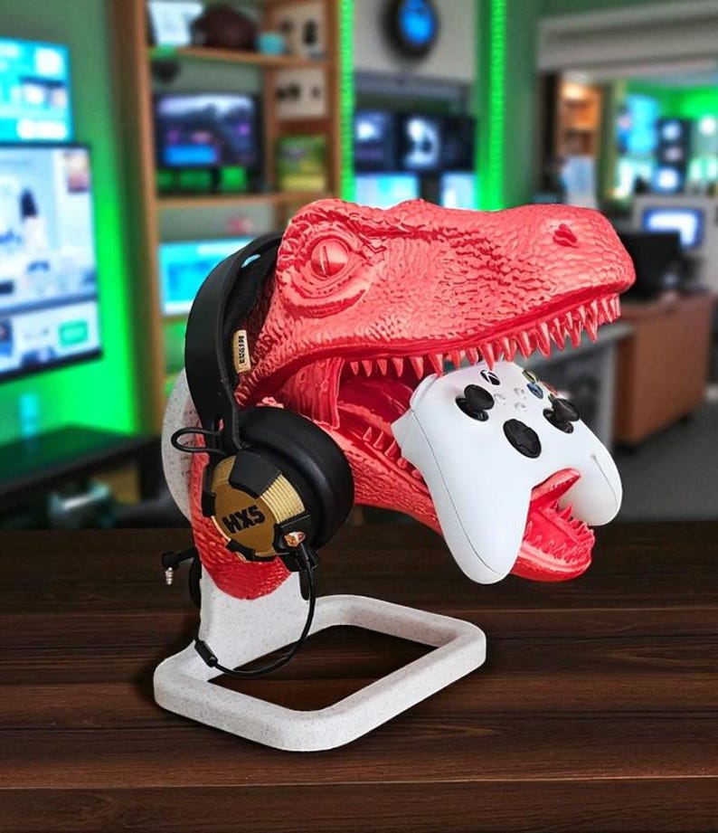 May include: A red dinosaur-shaped gaming headset and controller stand. The stand holds a black headset with gold accents and a white controller. The stand is on a white base and sits on a wooden surface.