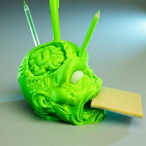 May include: A green, 3D-printed desk organizer shaped like a zombie head. The brain is visible inside the skull. It holds pens, pencils, and a stack of yellow sticky notes. The organizer is a unique and quirky desk accessory.
