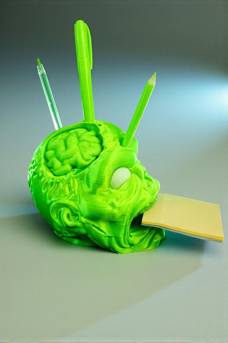 May include: A green, 3D-printed desk organizer shaped like a zombie head. The brain is visible inside the skull. It holds pens, pencils, and a stack of yellow sticky notes. The organizer is a unique and quirky desk accessory.